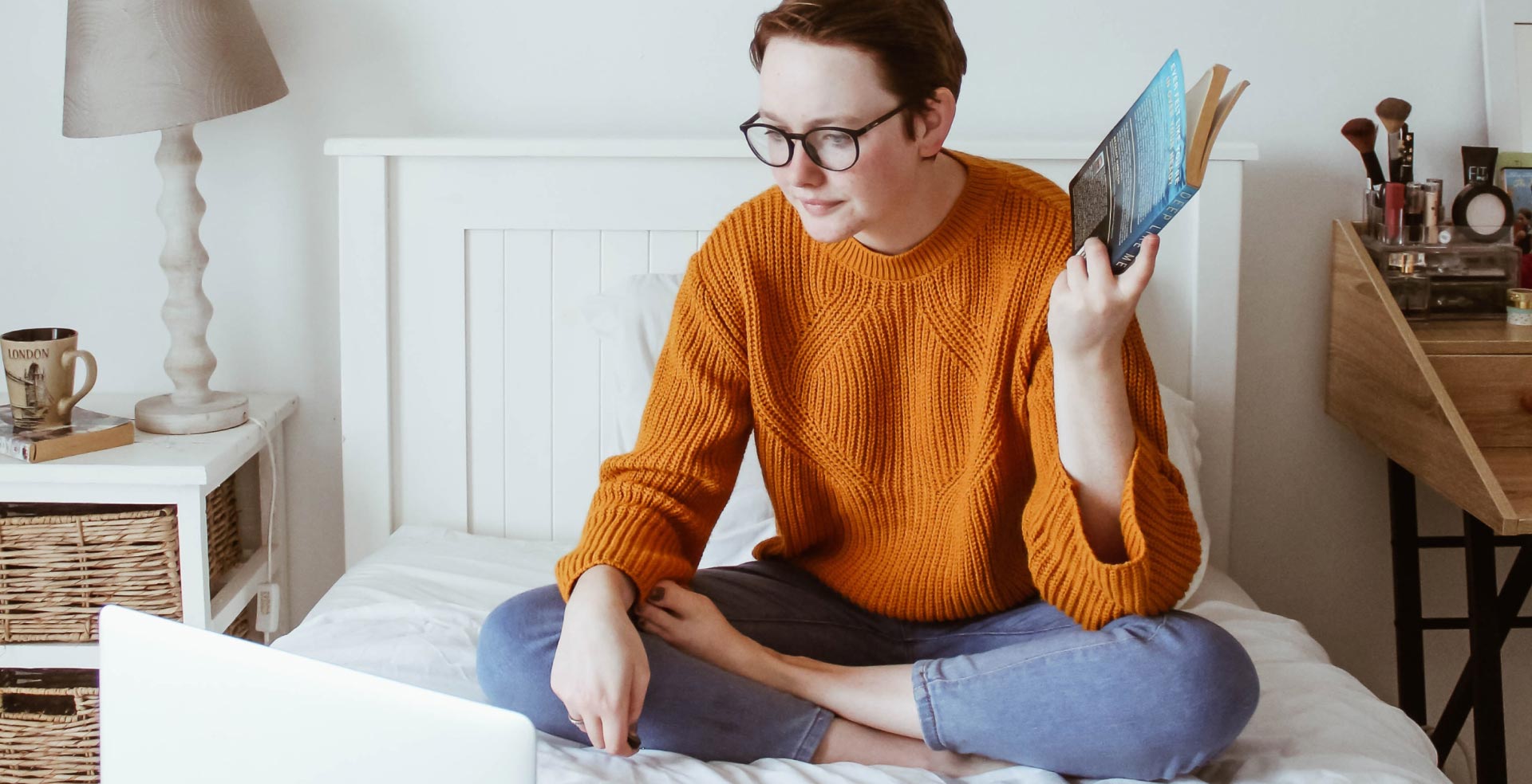 A woman sitting on her bed with a book in her hand looking at her laptop in front of her.