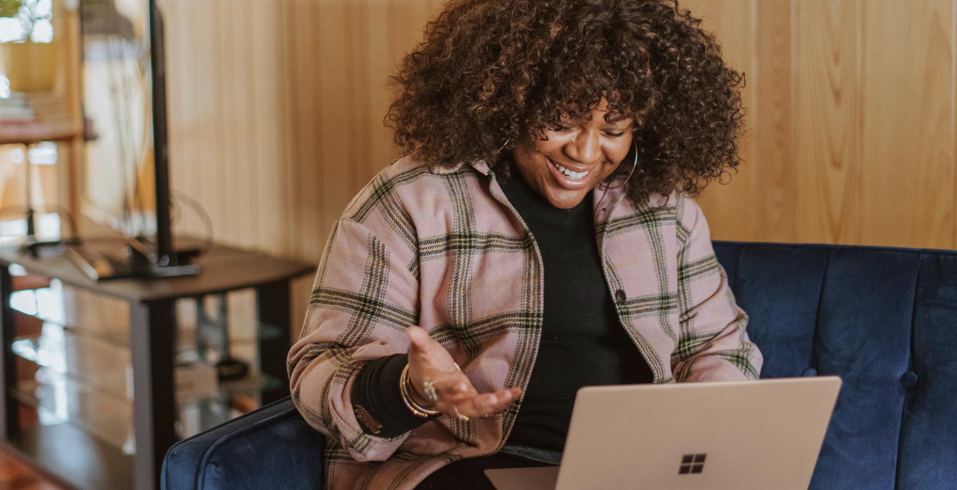 A woman sitting on a couch smiling while looking at her laptop.