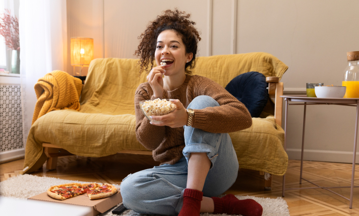 A woman sitting on a living room floor watching T.V and eating from a bowl of popcorn.