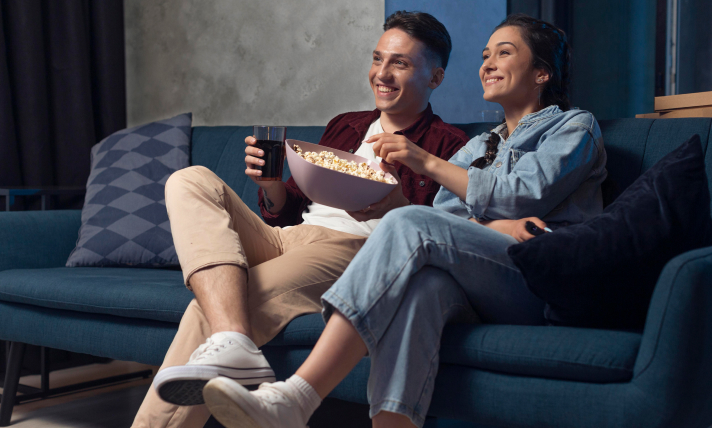 A man and a woman sitting on a couch watching T.V and eating from a bowl of popcorn.