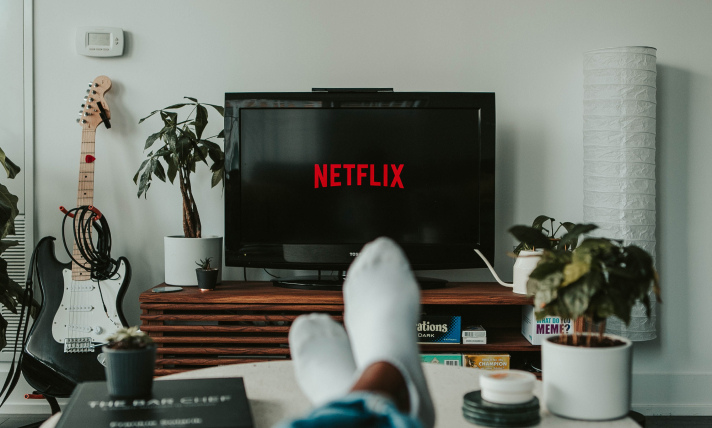 A T.V with the Netflix logo on the screen and a person's feet rested on a coffee table in the foreground.