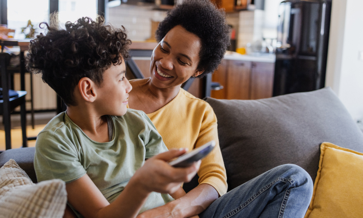 A woman and a little boy, who is holding a T.V remote control, sitting on a couch and smiling at each other.