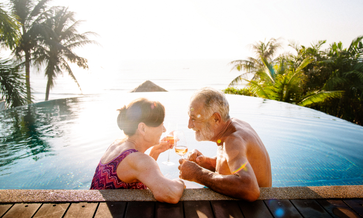 A man and a woman sitting in a pool in a tropical location smiling at each other.