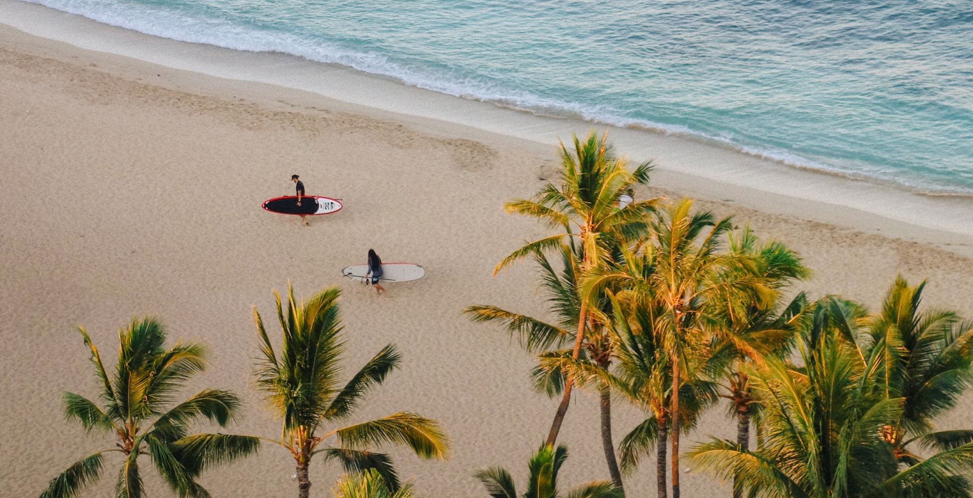 Aerial view of a beach in Hawaii with two people holding surf boards walking.