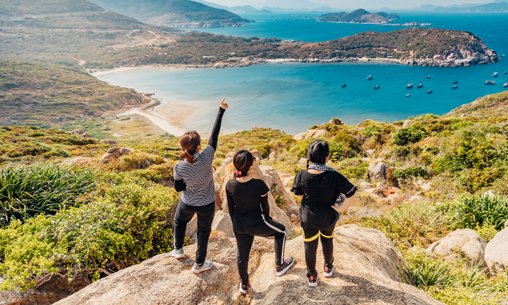 Three people standing on the edge of a cliff overlooking a cove with stunning blue water.