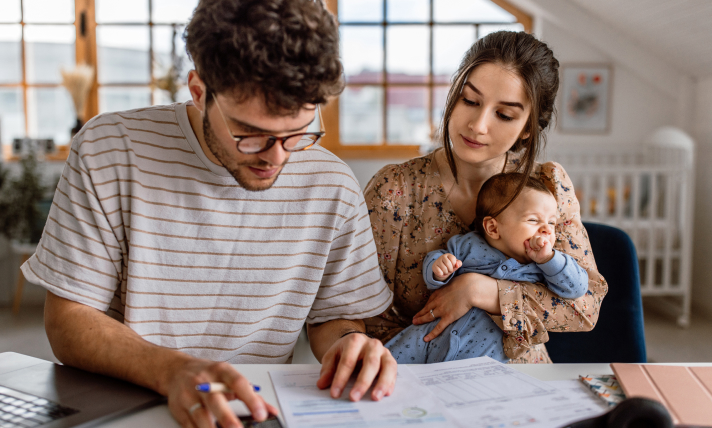 A man and a woman holding a baby reading a piece of paper on the table in front of them.