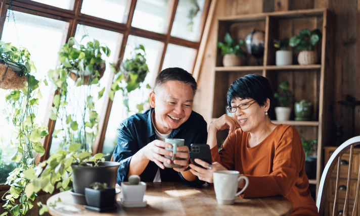 A man and a woman sitting at a table smiling while looking at a smartphone the woman is holding.