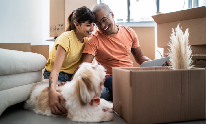 A man and a woman sitting on the floor of their living room next to their dog and a moving box.