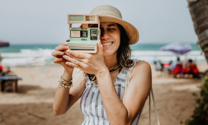 A woman on a beach taking a photo with a Polaroid camera.