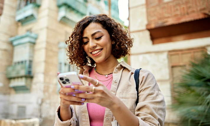 A woman walking down a street smiling while reading off of her smartphone.