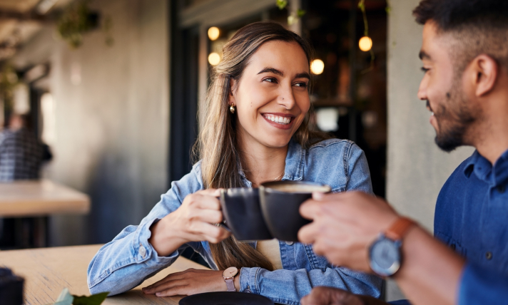 A man and a woman smiling while they clink two coffee mugs together.