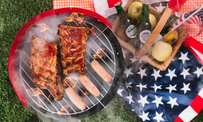 A barbeque grilling ribs beside a table set with Fourth of July decorations, apples, and popcorn.