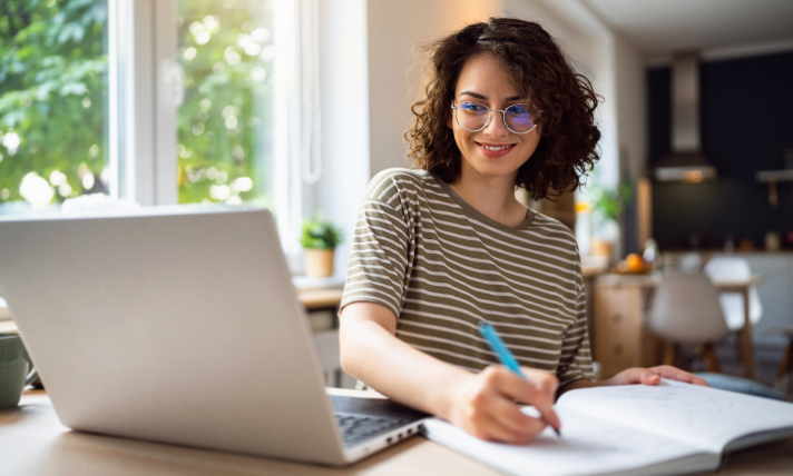 A woman writing on a notepad while looking at a laptop.