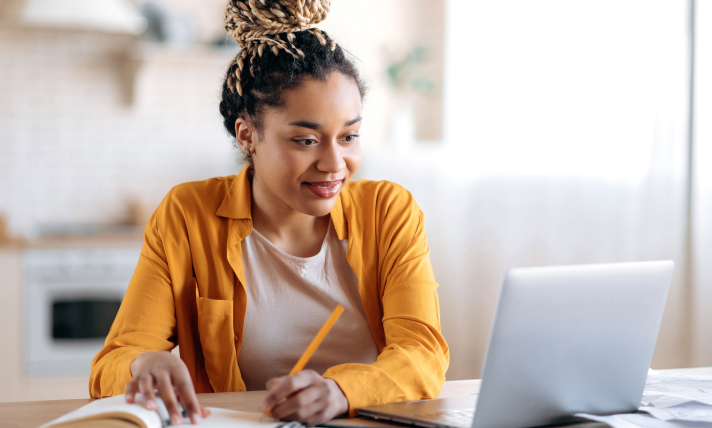 A woman looking at a laptop screen while writing in a notepad.