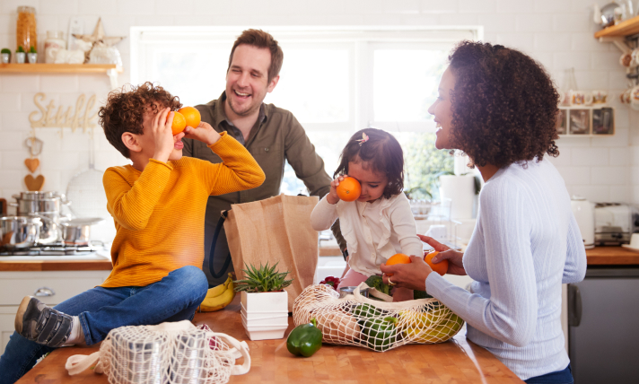 A man, woman, and two children smiling as they unpack groceries in a well-lit kitchen.