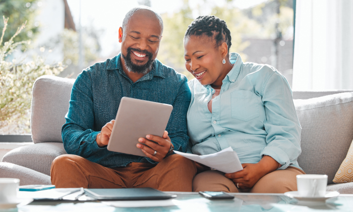 A man and a woman sitting on a couch and smiling at a computer tablet.