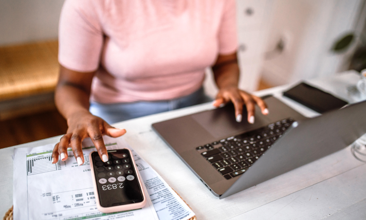 A woman sitting at a desk typing on her computer with one hand and her phone calculator with her other hand.