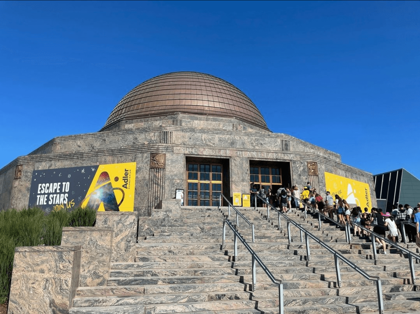 The outside of the Adler Planetarium with people lined up on the steps on a summer day with blue sky.