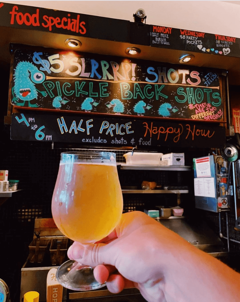 Small glass of beer being held up in front of a chalkboard advertising drink specials.