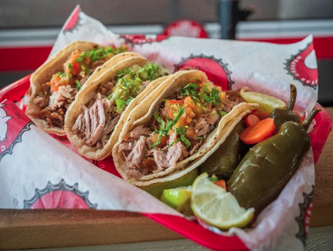 Three tacos filled with various meats and vegetables in a red takeaway container.